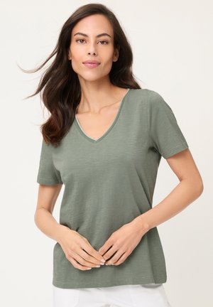 Frau mit langen dunklen Haaren, die ein salbeigrünes V-Ausschnitt-Kurzarmshirt und weiße Hose trägt, steht mit verschränkten Händen.