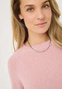 Helles rosa Sweatshirt aus weichem Stoff mit einem runden Ausschnitt. Eine silberne Halskette hat ein kreisförmiges Element und verlängerte Glieder.