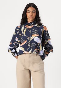 Blusa floreale blu navy con maniche a palloncino, colletto arricciato e vivaci motivi floreali in arancione, rosa e bianco, abbinata a pantaloni beige.