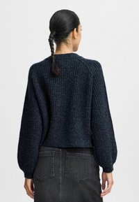 Tmavě námořnický svetr se žebrovanou strukturou, kulatým výstřihem a nabíranými rukávy. Nosený přes šedou džínsovou sukni s kapsami na zadní straně.