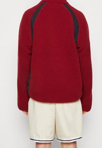 Rote Fleecejacke mit strukturiertem Material, schwarzen Seitenteilen und hohem Kragen, kombiniert mit beigen Shorts.