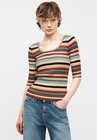 Γυναίκα με κοντά κυματιστά μαλλιά που φοράει μια ριμπ πλεκτή μπλούζα με πολύχρωμες ρίγες και μανίκια μέχρι τον αγκώνα, καθώς και μπλε τζιν ψηλόμεσο με το χέρι στην τσέπη.