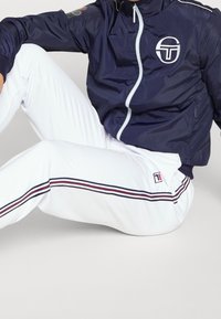 Námořnická větrovka s bílým logem, má přední zip a elastické manžety; spárována s bílými tepláky, doplněnými červenými a modrými pruhy.