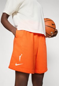 Oransje idrettsshorts laget av mykt stoff, med en hvit Nike-logo og en silhuett av en spiller. Sammen med en hvit t-skjorte med korte ermer.