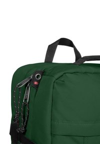 Vihreä kangasreppu, jossa on teksturoidumpi pinta, pääkahva ja mustat yksityiskohdat, kuten vetoketju ja naru. Sivutasku näkyvissä.