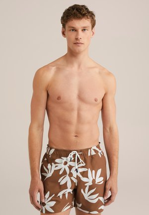 Braune Badeshorts mit großen weißen Blumenmustern, elastischem Bund und Kordelzug. Das Model posiert vor einem neutralen Hintergrund.