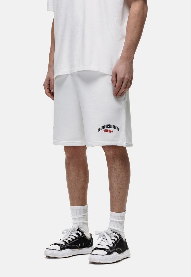 Pantaloni corti bianchi in cotone con vestibilità ampia, caratterizzati da ricami neri e rossi con la scritta "GOODFORNOTHING". Abbinati a sneaker nere e calzini bianchi.