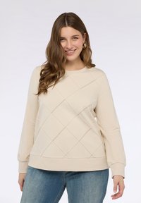 Beige mikina s prošívaným diamantovým vzorem, dlouhými rukávy a ribovanými manžetami, v kombinaci se světle modrými džíny. Hladká textury látky.