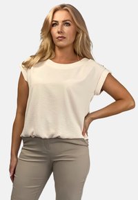 Jasno beżowa bluzka z krótkim rękawem, okrągłym dekoltem i podwiniętymi mankietami. Gładka tekstura, luźny krój i subtelny połysk. Zestawiona z jasno beżowymi spodniami.