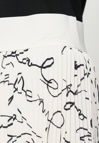 Fehér anyagból készült pliszírozott szoknya fekete absztrakt mintákkal, széles rugalmas derékpánttal, melyen világos szállal hímzett "Karl" felirat található.