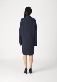 Navy ribbed gebreid jurk met een brede kraag, lange mouwen en een oversized fit, gedragen met zwarte hoge hakken.