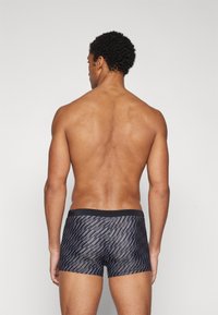 Mann med krøllete hår som står vendt bort, iført svart og grå mønstrede boxershorts mot en enkel lys bakgrunn.