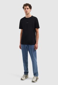 Man met krullend haar, gekleed in een zwart "PURE PATH" T-shirt, blauwe jeans en witte sneakers, staand tegen een effen lichtgrijze achtergrond.