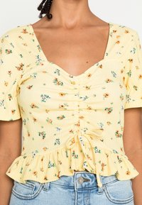 Donna che indossa un top arricciato floreale giallo con maniche corte e orlo a volant, abbinato a jeans di denim azzurro chiaro.