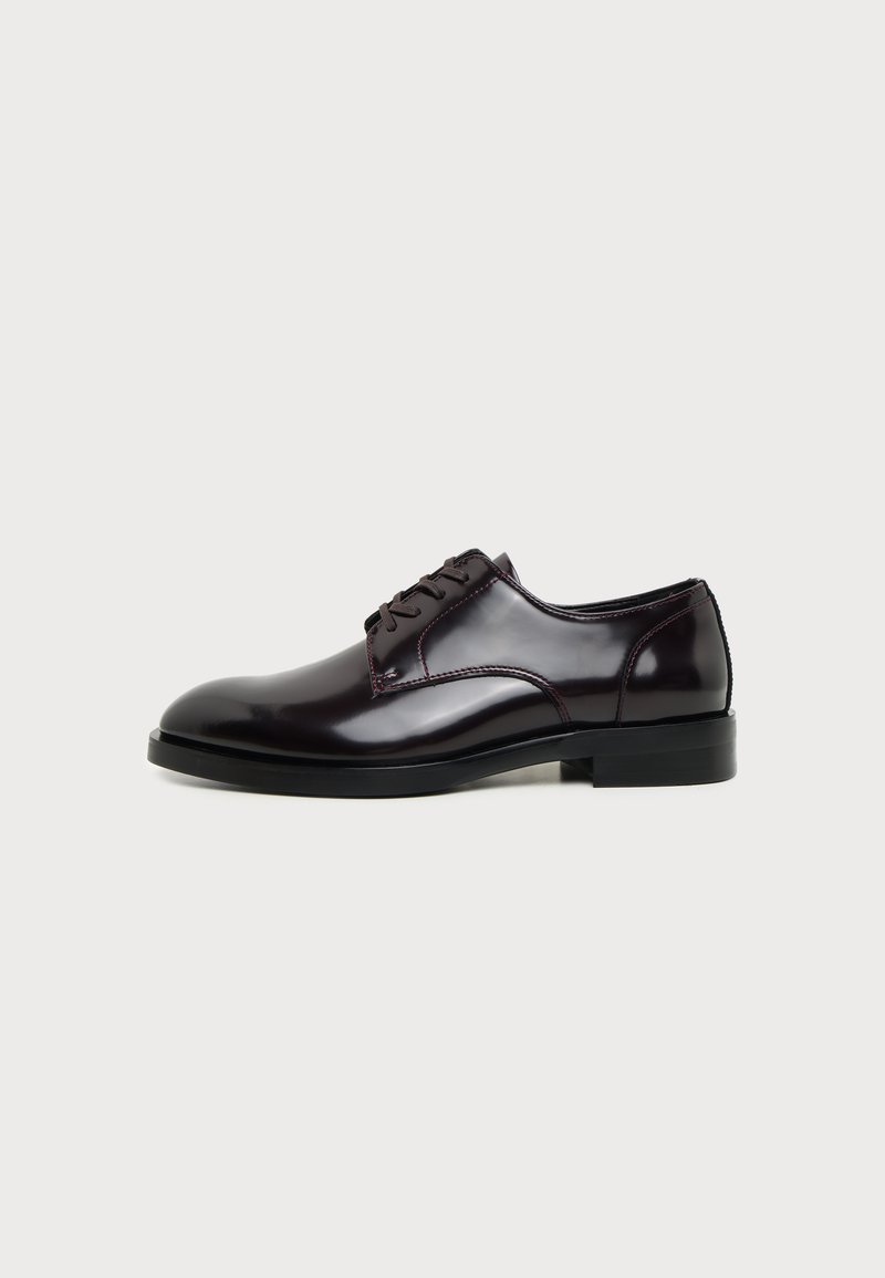 Chaussure habillée pour homme en cuir noir brillant avec lacets à l’avant et petit talon, montrée de profil sur fond clair.