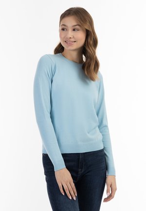 Giovane donna con capelli castani ondulati che indossa un maglione azzurro chiaro e jeans scuri, sorridente e che guarda leggermente di lato.