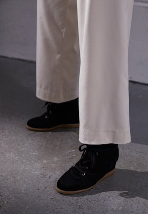 Osoba ubrana w kremowe spodnie o szerokich nogawkach i czarne zamszowe sneakersy na koturnie stojąca na szarej betonowej podłodze.