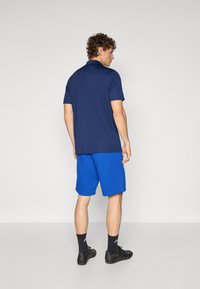 Polo azul-marinho com barra reta, combinado com calções azuis até ao joelho. Modelo a usar sapatos e meias pretos com marcação branca.