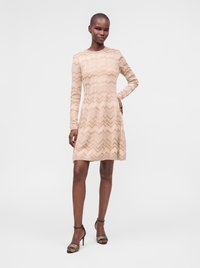 Femme portant une robe beige à manches longues avec un motif zigzag et des talons hauts ouverts noirs, debout devant un fond blanc uni.