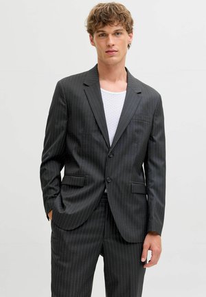 Ung mann kledd i en mørkegrå dressjakke med tynne striper og matchende bukser over en hvit teksturert skjorte, står foran en ensfarget bakgrunn.