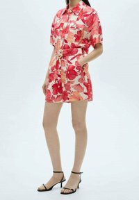 Sukienka koszulowa w kwiaty w kolorze czerwonym i kremowym, z krótkimi rękawami, z guzikiem z przodu, z paskiem w talii, z bocznymi kieszeniami i luźnym krojem. Czarne sandały na ramiączkach.