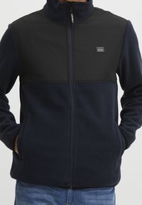 Man draagt een marineblauwe fleecejas met een zwarte schouderpas, een voorsluiting met rits, steekzakken en een klein logo op de borst, gecombineerd met een blauwe spijkerbroek.