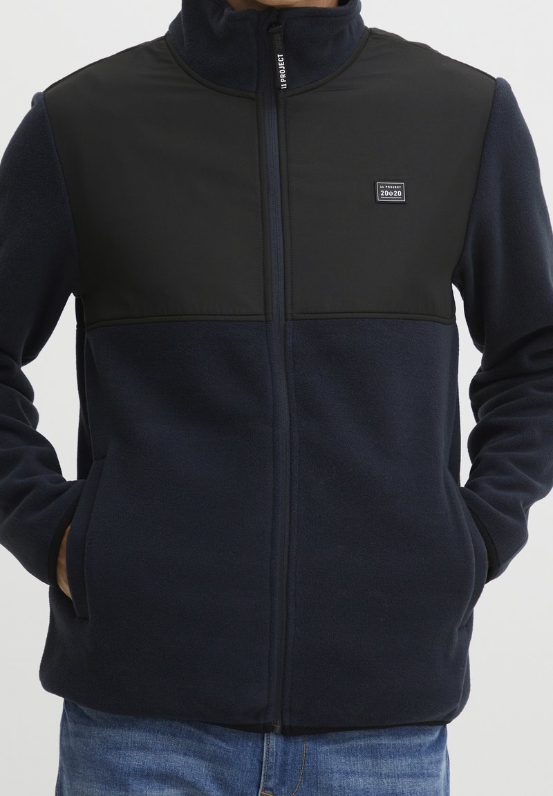 Man draagt een marineblauwe fleecejas met een zwarte schouderpas, een voorsluiting met rits, steekzakken en een klein logo op de borst, gecombineerd met een blauwe spijkerbroek.