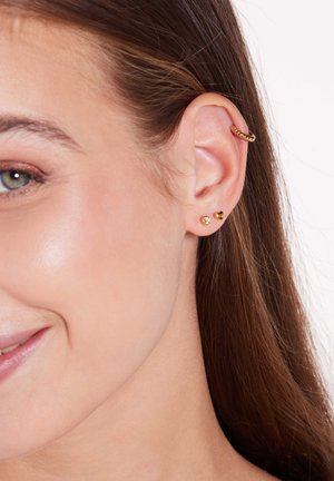 Gouden oorbellen tentoongesteld: een gedraaide oorring, twee studs - één met een heldere steen en één in de vorm van een hart. De huidtint complimenteert de sieraden en versterkt de kenmerken.