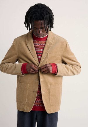 Beige Cordjacke mit Revers und zwei vorderen Taschen, getragen über einem rot gemusterten Pullover mit geripptem Ausschnitt.