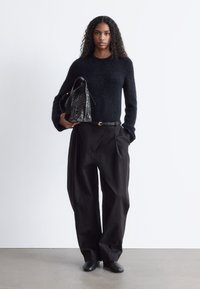 Schwarzer, flauschiger Pullover, hochgeschnittene schwarze Hose mit Falten, schwarze Tasche mit strukturiertem Finish, schwarze Slipper, neutraler Hintergrund.