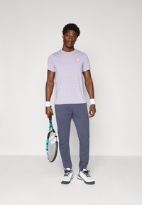 Camiseta deportiva de manga corta color lila claro con rayas horizontales, combinada con pantalones de chándal azul marino y zapatillas de tenis blancas, sosteniendo una raqueta de tenis.