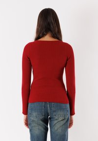 Top lungo rosso a coste con maniche lunghe, aderente in vita, abbinato a jeans in denim blu a vita alta. Il top presenta un ampio scollo e un tessuto strutturato.