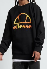 Person som bär svart sweatshirt med orange-gul gradient "ellesse" logotyp, vit skjorta under och mörka byxor, står framför en enfärgad bakgrund.
