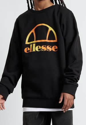 Personne portant un sweat à capuche noir avec un logo "ellesse" en dégradé orange-jaune, une chemise blanche en dessous et un pantalon sombre, se tenant devant un fond uni.