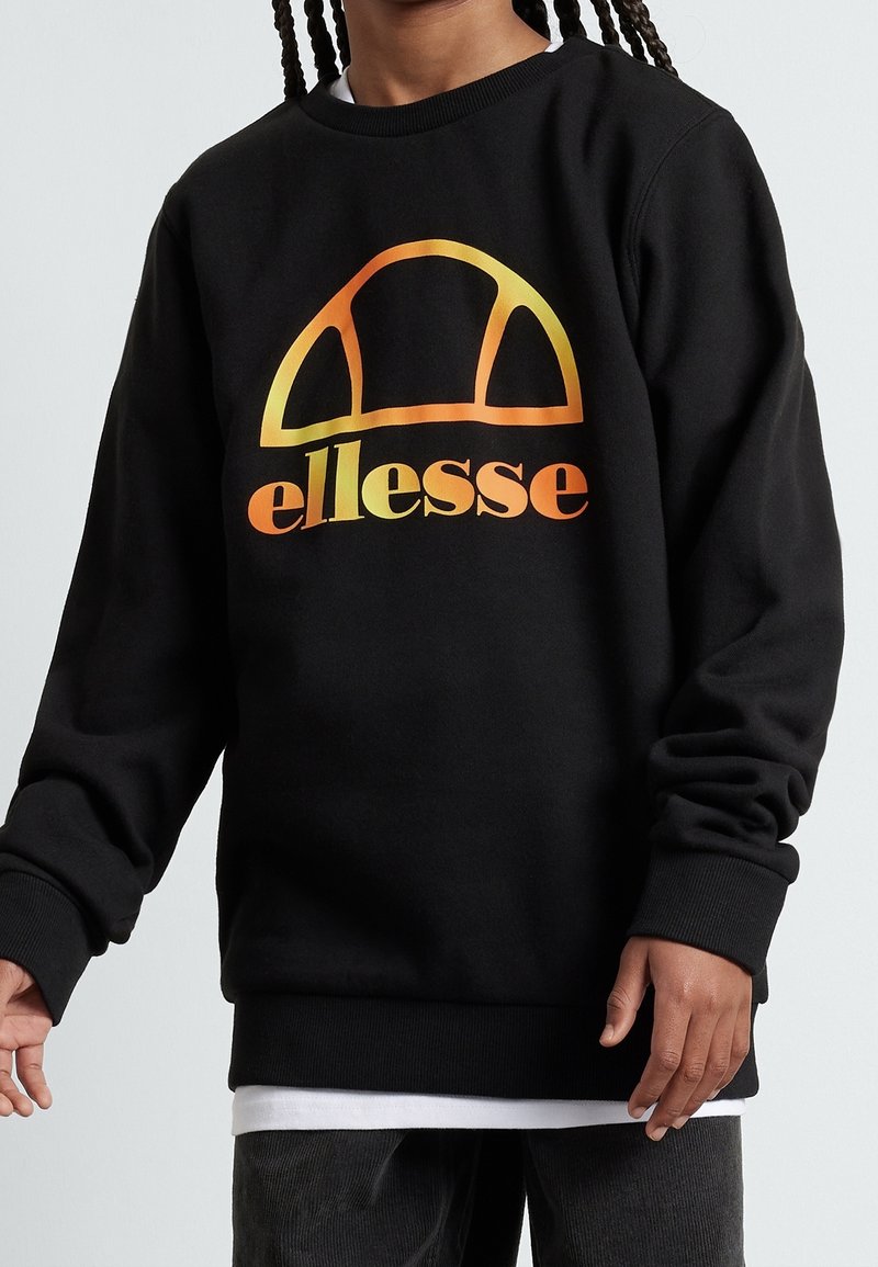 Person som bär svart sweatshirt med orange-gul gradient "ellesse" logotyp, vit skjorta under och mörka byxor, står framför en enfärgad bakgrund.
