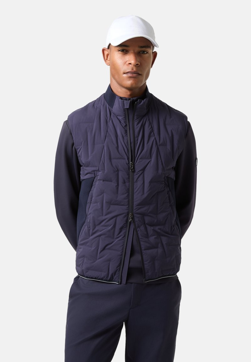 Navy padded vest with quilted texture, full zip, high neck, and side pockets, paired with a dark outfit and a white cap.