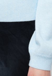 Gaiši zils adīts džemperis ar ribotām piedurknēm, apvienots ar melniem zamša biksēm. Ievērojams tekstūras kontrasts starp materiāliem.