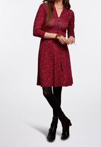 Žena oblečená v červených patterned košilových šatech s třičtvrtinovými rukávy, černými punčochami a černými kotníkovými botami, stojící se zkříženými nohama.