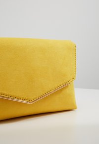 Pochette enveloppe en daim jaune avec bordure dorée, posée sur une surface blanche devant un fond gris clair.