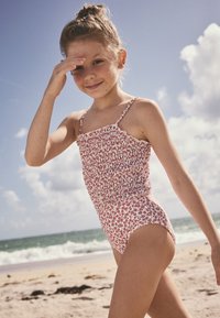 Maillot de bain une pièce fleuri blanc à motifs rouges, avec un corsage smocké et des bretelles fines réglables, porté sur une plage de sable.