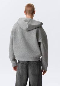 Person mit kurzen Haaren, der einen grauen Kapuzenpullover und dunkelgraue Jeans trägt, von hinten vor einem schlichten Hintergrund zu sehen.