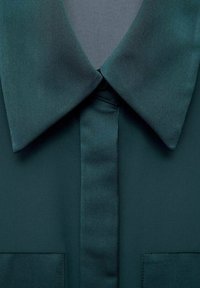 Dark green blouse featuring a large collar, button placket, and two chest pockets. The fabric has a smooth, shiny texture.