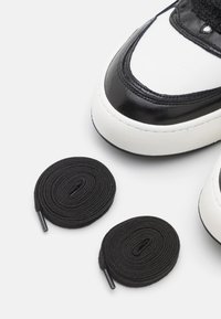 Zbliżenie na czubek czarno-białego trampka z dwoma zwiniętymi czarnymi sznurowadłami na białym tle.