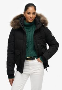 Sort pufjakke med hætte besat med pels, lynlås og tryknapper, to frontlommer, båret over en grøn cable-knit sweater og hvide bukser.
