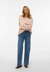 Frau steht mit verschränkten Armen in einem hellrosa schulterfreien Pullover, hochgeschnittenen blauen Jeans, einem schwarzen Gürtel und braunen Schuhen vor weißem Hintergrund.