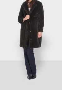 Mörkgrön pälsjacka med bred krage, prydd med guldknappar fram, bärs över blå jeans och svarta ankelboots.