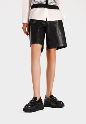 Pantaloni di pelle nera con una texture liscia, caratterizzati da un taglio dritto e un design di lunghezza media, abbinati a scarpe nere chunky.