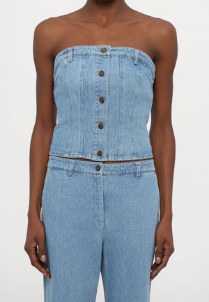 Vrouw draagt lichtblauwe strapless denim top met knopen en bijpassende hooggetailleerde denim broek tegen een effen achtergrond.