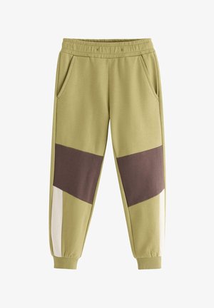 Smilende ung dreng stående med hænderne i lommerne, iført en farveblokeret sweatshirt i grøn, brun og creme og matchende bukser med sneakers.