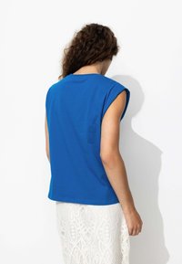 T-shirt sans manches bleu avec un col rond, fabriqué en coton doux. Présente un design simple et une coupe légèrement décontractée au dos.